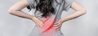 Kobieta z czarnymi włosami w szarej koszulce stoi tyłem, trzymając ręce na dolnej części pleców, która jest czerwona z powodu bólu. Ilustracja dotycząca rwy kulszowej.