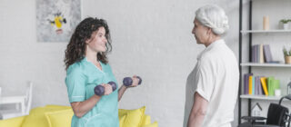 Młoda kobieta wykonuje ćwiczenia z fizjoterapeutą, trzymając dwa hantle w rękach, uśmiechnięta i pełna energii podczas sesji rehabilitacyjnej.